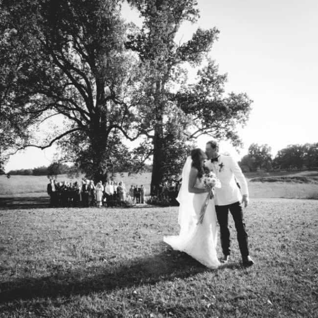 Bride and groom walking hand in hand with guests in the background, monochrome outdoor wedding scene.
