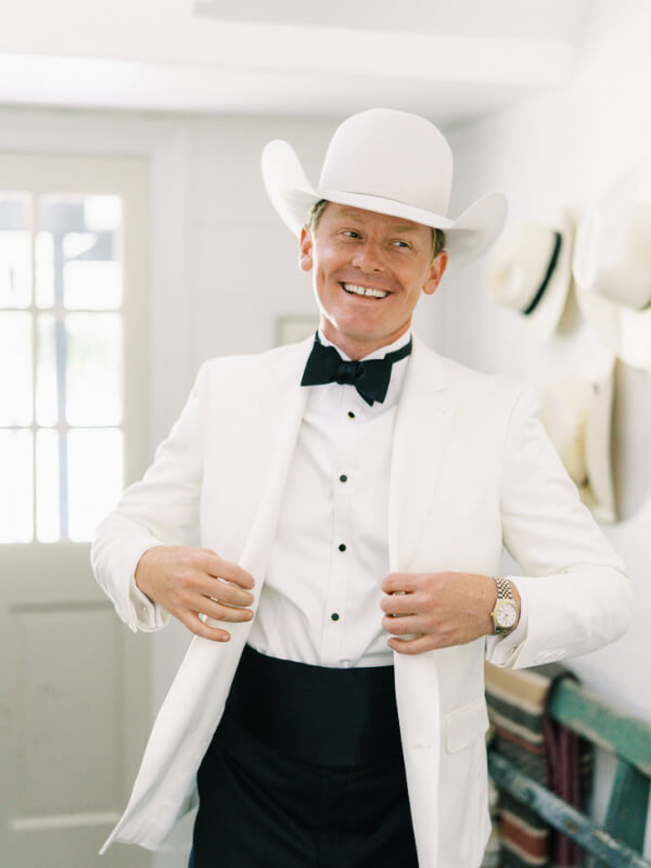A man in a white suit and cowboy hat smiling indoors.