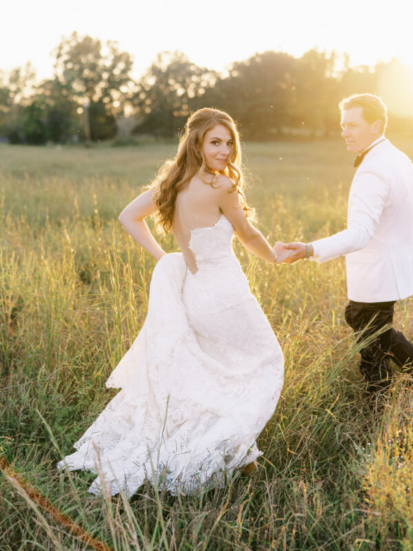 A bride in a lace gown holding hands with a groom in a white jacket in a sunlit field.