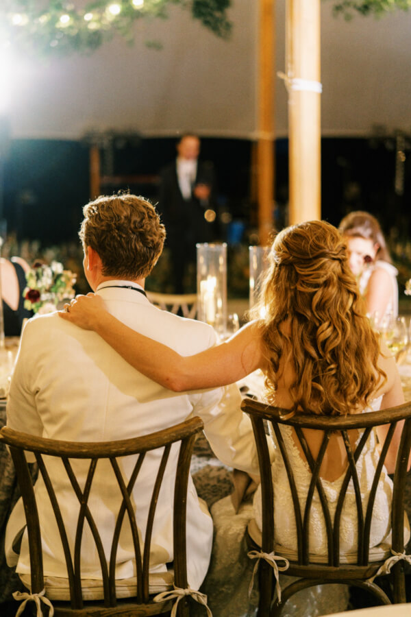 A couple in formal attire seated back-to-back at an event, with a speaker in the background.