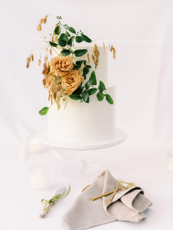 Elegant two-tiered white wedding cake adorned with dried flowers and greenery on a pedestal, accompanied by neatly arranged cutlery on a linen napkin.