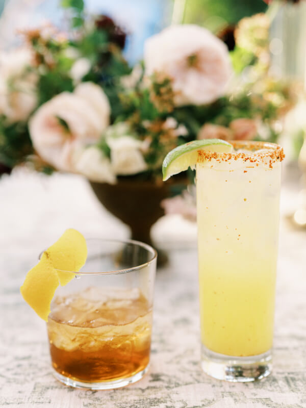 Two cocktails on a table with a floral centerpiece in the background.