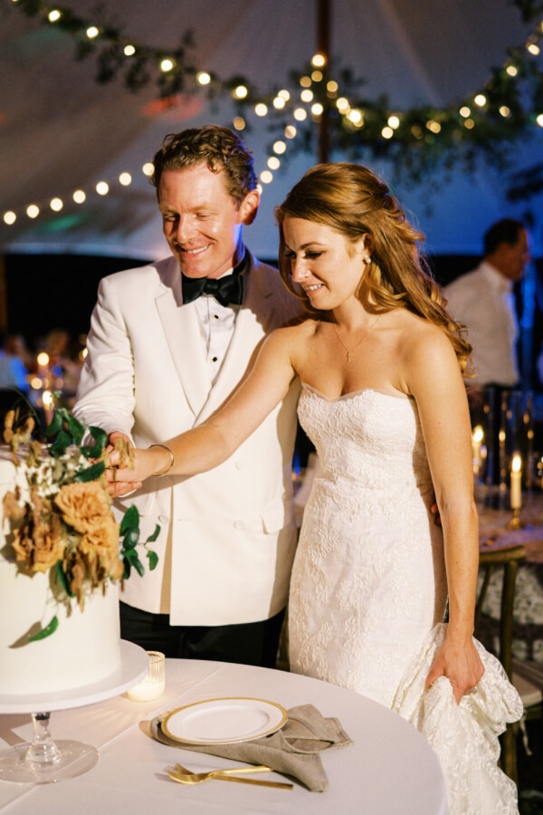 A bride and groom cutting their wedding cake under a tent adorned with string lights.