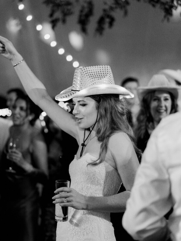 A bride in a white dress and a straw cowboy hat dancing at a wedding.