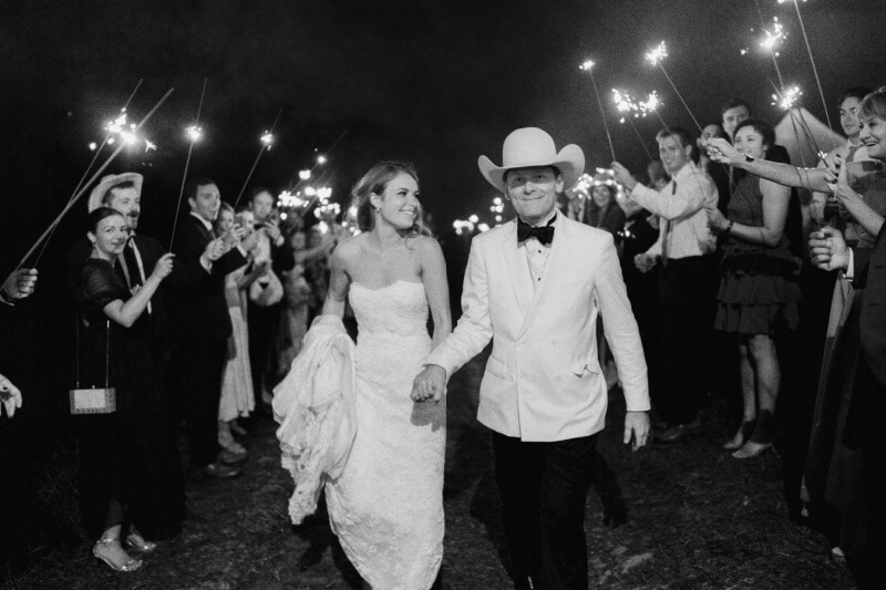 A bride and groom walk hand in hand through a tunnel of guests holding sparklers.