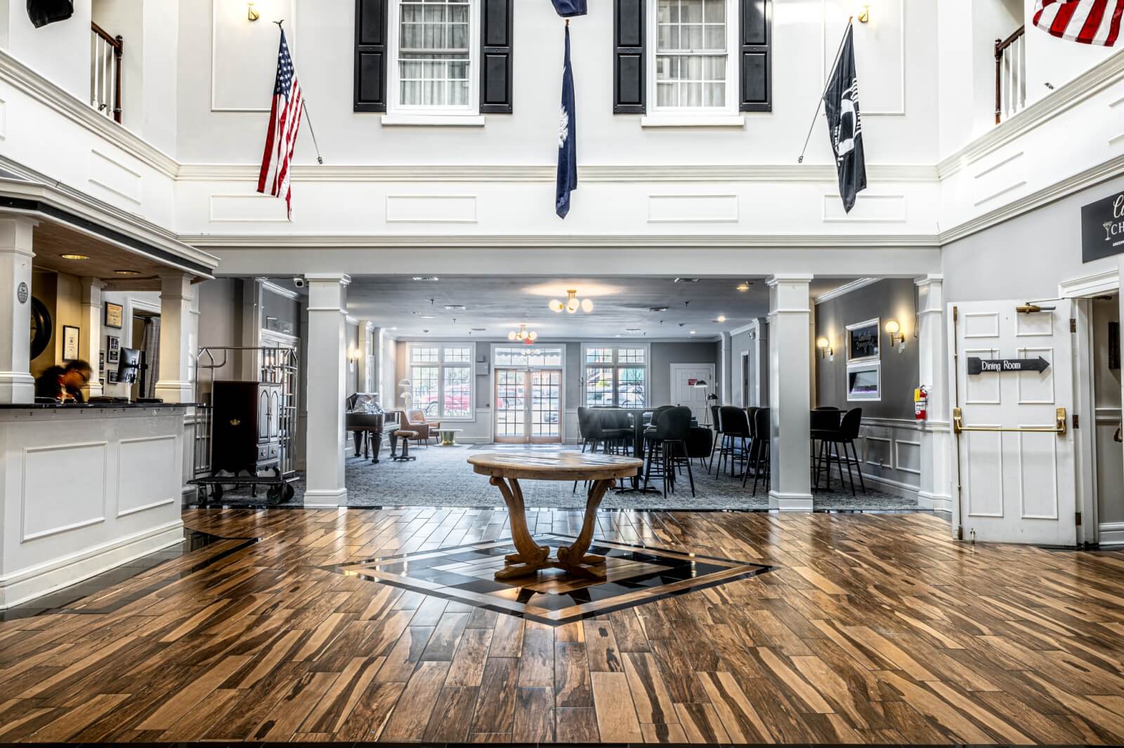 The bright hotel lobby, ideal for retreats in South Carolina, features a wooden floor, a round table at the center, and flags on the balcony above. The reception desk is on the left, a seating area graces the background, and double doors invite you in on the right.