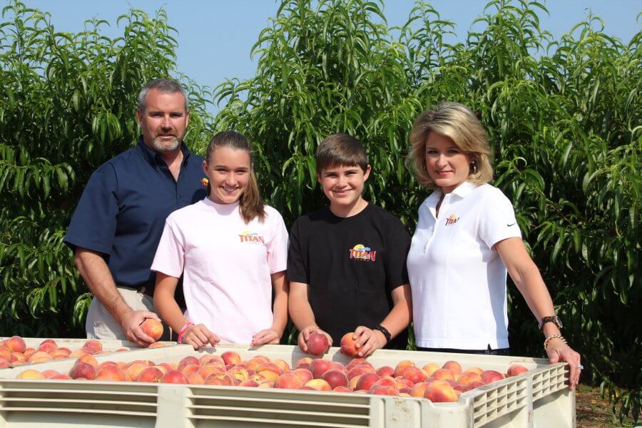A family of four standing behind a large crate of peaches at Titan Farms orchard, smiling at the camera.