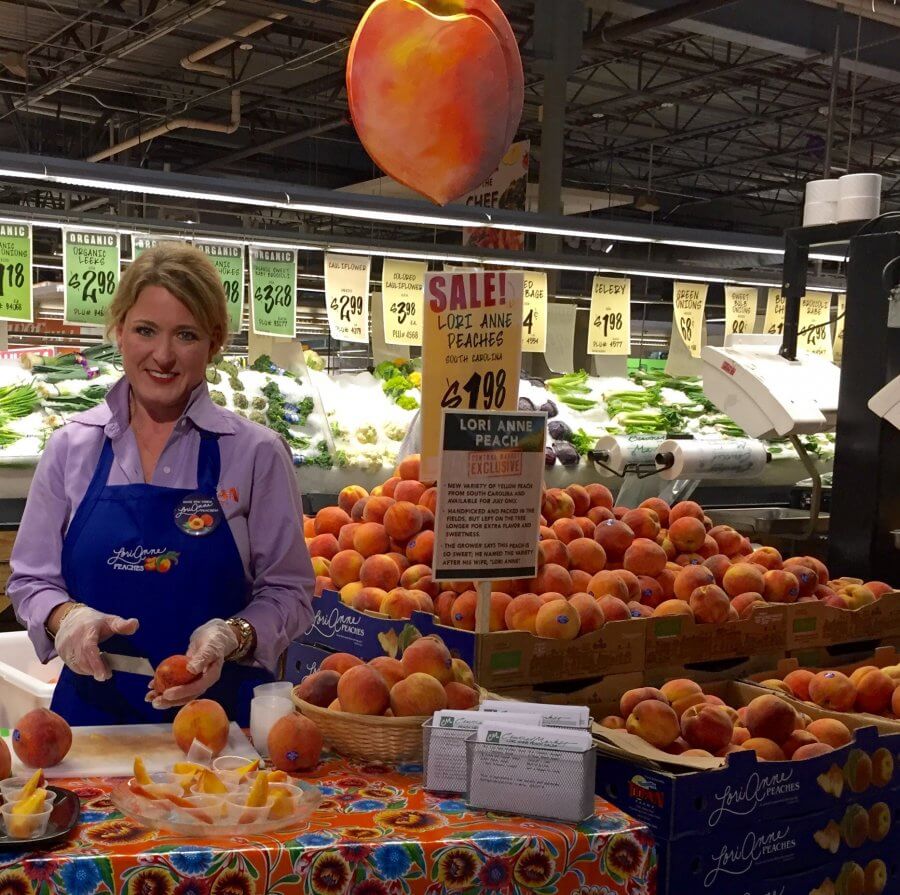 A woman in a blue apron stands behind Titan Farms' grocery store's peach display, next to a "sale!" sign, with bins of fresh peaches around her.