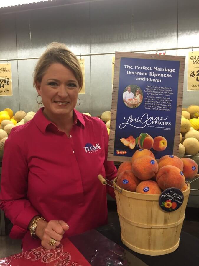 Woman in a red shirt standing beside a display of peaches in a grocery store, promoting Titan Farms peaches with a sign.