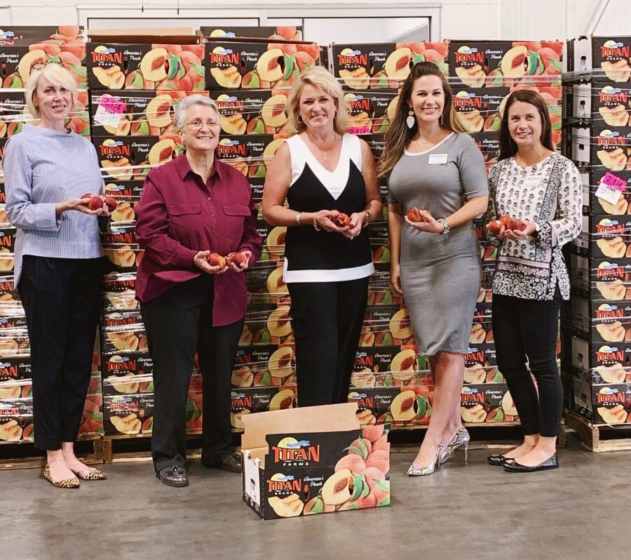 Five women standing in a warehouse owned by Titan Farms, holding peaches in front of stacked boxes of fruit.