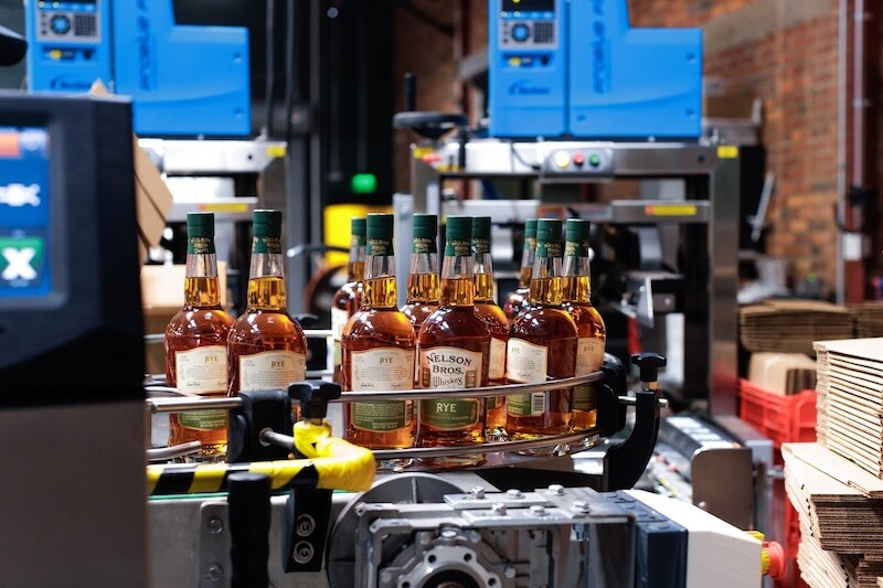 Bottles of rye whiskey being conveyed and processed in a factory, with blue machinery and brick walls visible in the background.