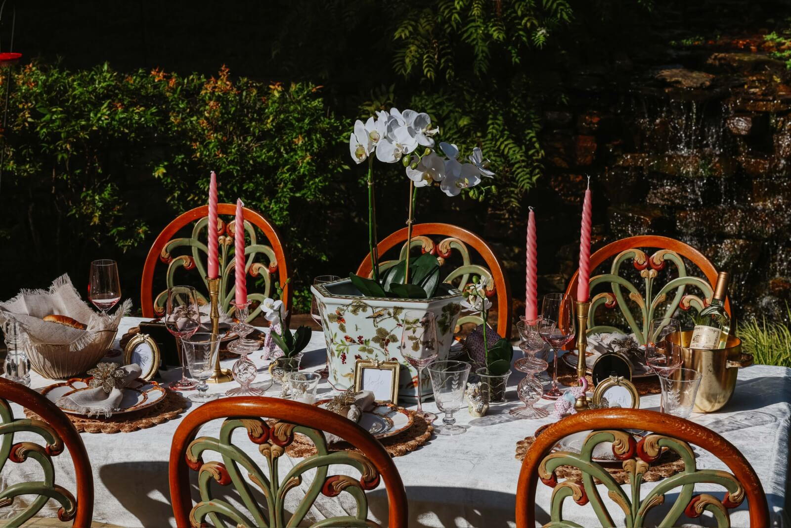An elegant outdoor dining setup features a white tablecloth, ornate chairs, pink candles, wine glasses, and a centerpiece of white orchids. With the soothing sounds of a small waterfall in the background, it’s as inviting as discovering the best gift shops in Nashville.