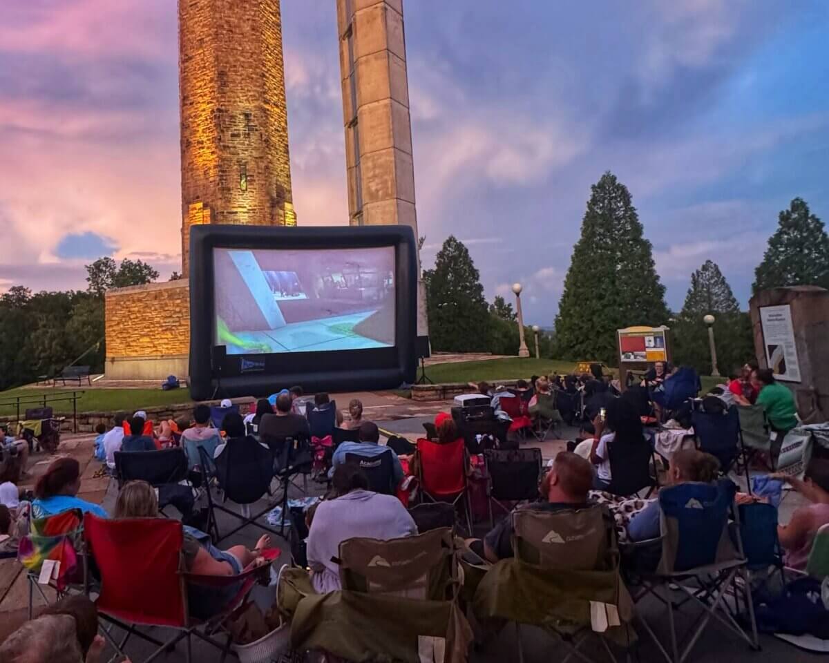 People sit in lawn chairs watching an outdoor Birmingham Movies screening projected on a large inflatable screen at dusk, with tall stone towers and trees in the background.