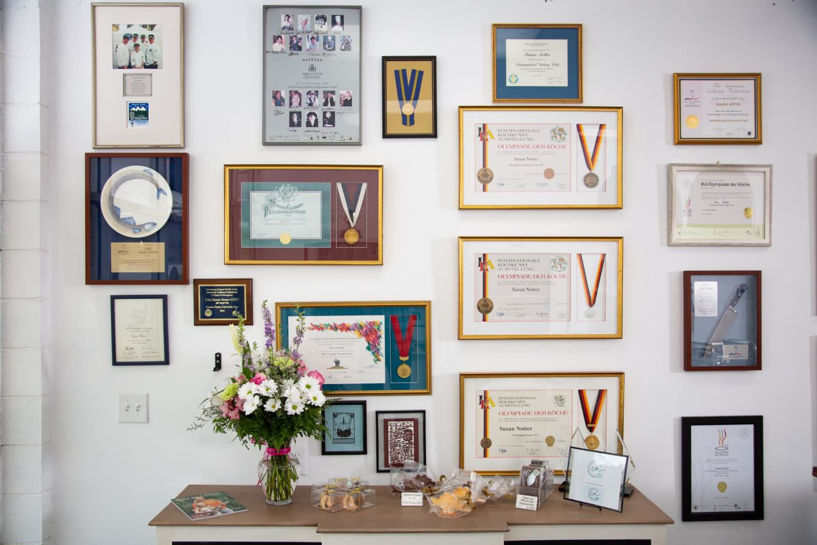 A wall displays numerous framed awards and certificates, with medals hanging on some. Below, a table holds a bouquet of flowers, cookies in plastic bags, and various small framed items celebrating the achievements of a world-renowned pastry chef.