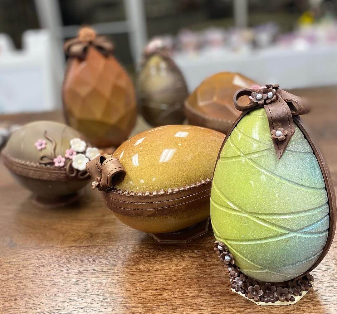 A collection of large, intricately decorated Easter eggs made of chocolate, some adorned with ribbons and floral details by Elizabeth Moore Chocolates, displayed on a wooden surface.
