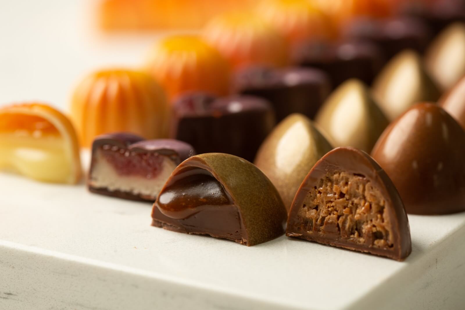 An assortment of specialty goumet chocolates from Elizabeth Moore Chocolates are lined up with the first row of four chocolates cut in half to reveal varied textures and fillings inside.