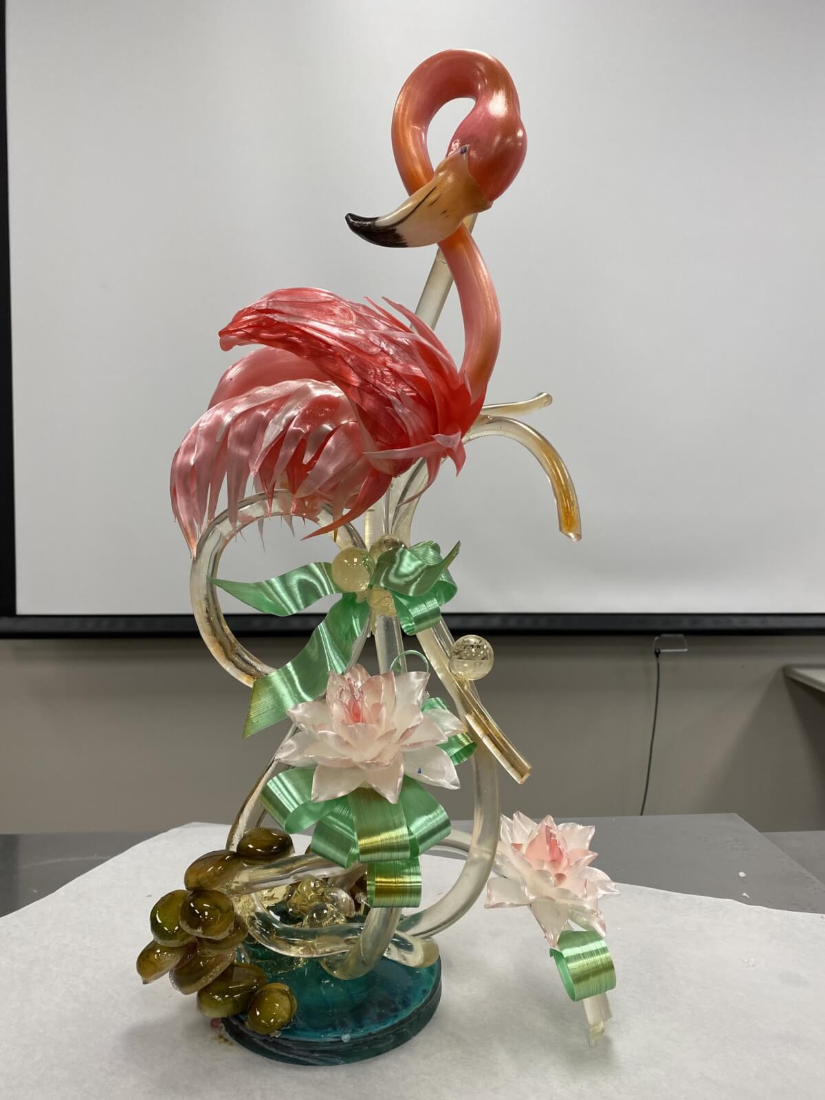 A sugar sculpture of a pink flamingo balanced on one leg, accompanied by green ribbon-like swirls and pink flowers on a white background from renowned Pastry Chef Susan Notter of Elizabeth Moore Chocolates.