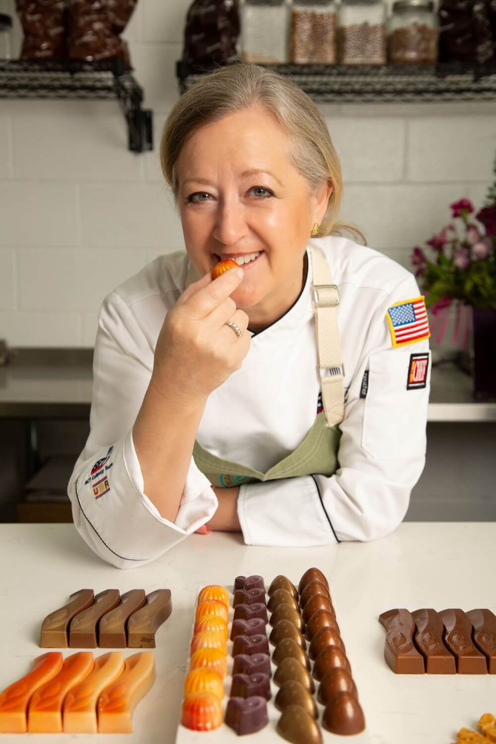 Pastry Chef Susan Notter in a white uniform enjoys a chocolate truffle in a kitchen setting, with multiple rows of Elizabeth Moore Chocolates neatly arranged on the counter. Shelves with jars are visible in the background.