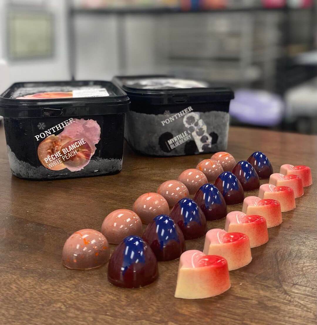A display of assorted colorful chocolate bonbons from Elizabeth Moore Chocolates sits in front of two tubs of fruit yogurt on a table, showcasing the artistry of Pastry Chef Susan Notter.