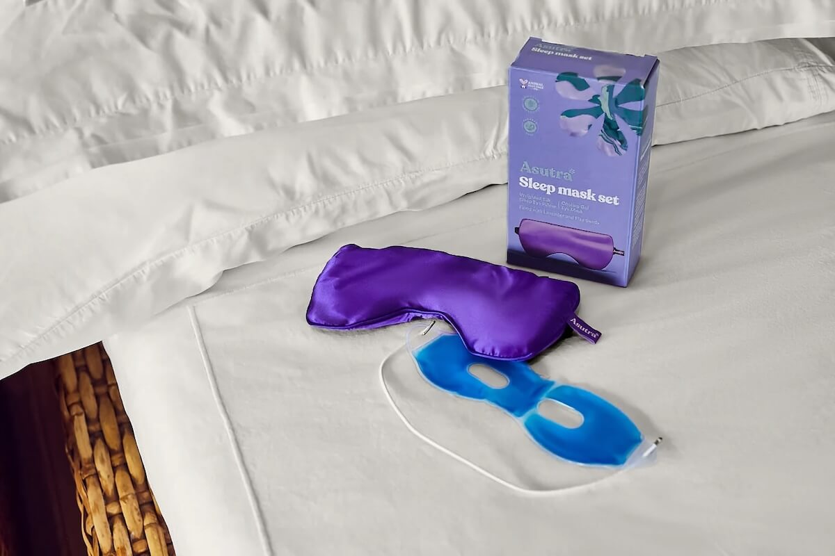 A purple sleep mask and a blue gel eye mask with its packaging box on a white bed sheet.