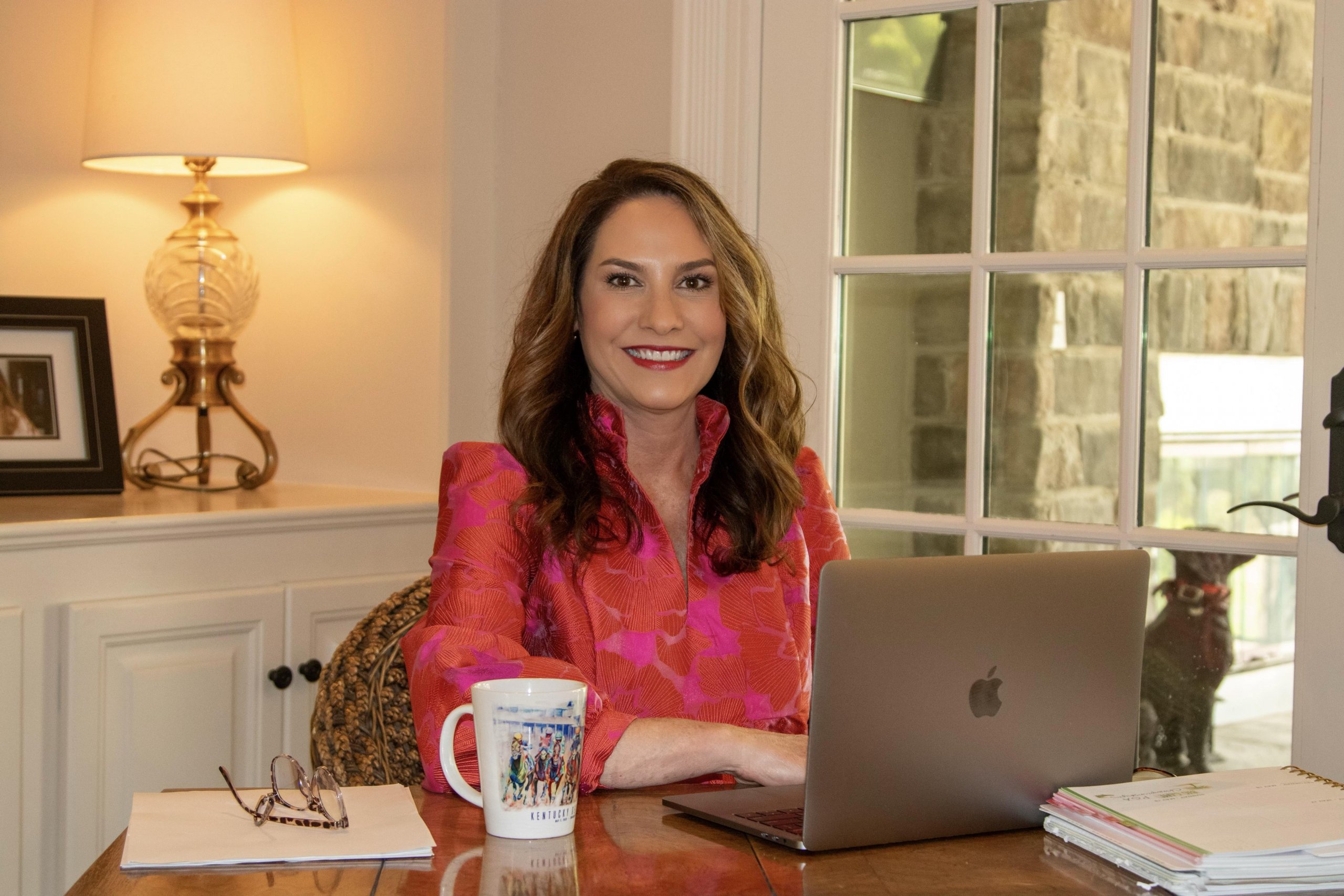 A woman smiling at the camera, seated at a desk with a laptop, mug, and documents, in a well-lit room with stone walls and a window.