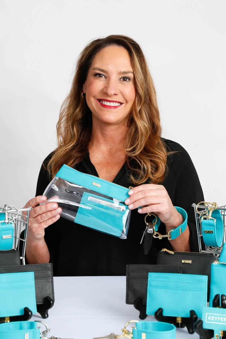 A smiling woman holding a clear, turquoise-trimmed pouch and standing in front of a display with similar items.