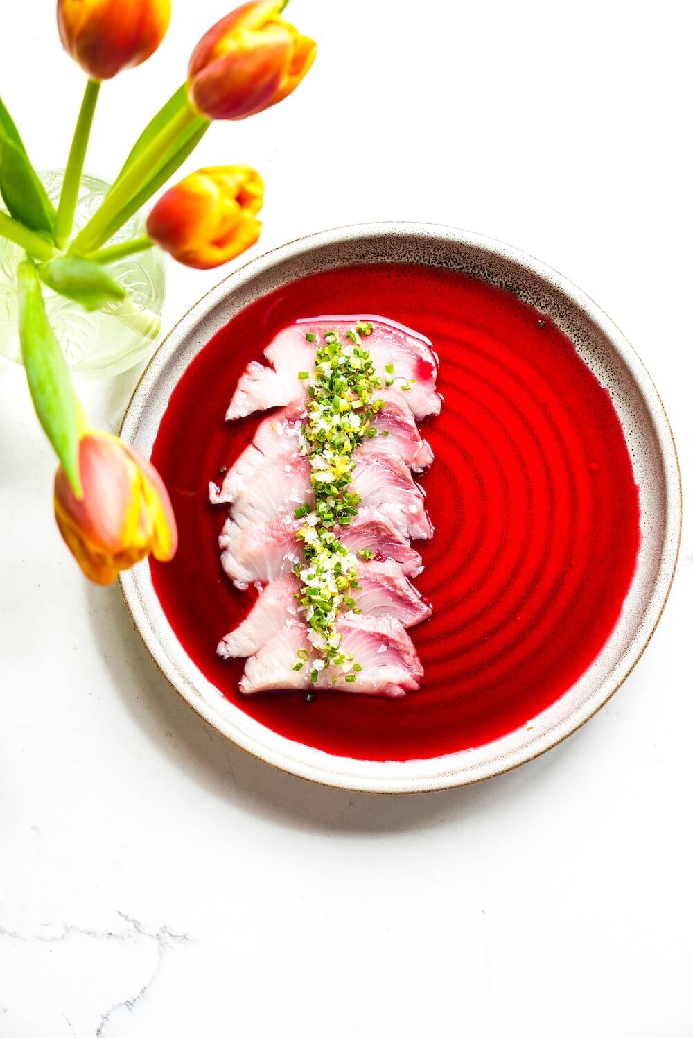 A plate of thinly sliced fish topped with green garnish is served in a vibrant red sauce, with a vase of orange and yellow tulips beside it.
