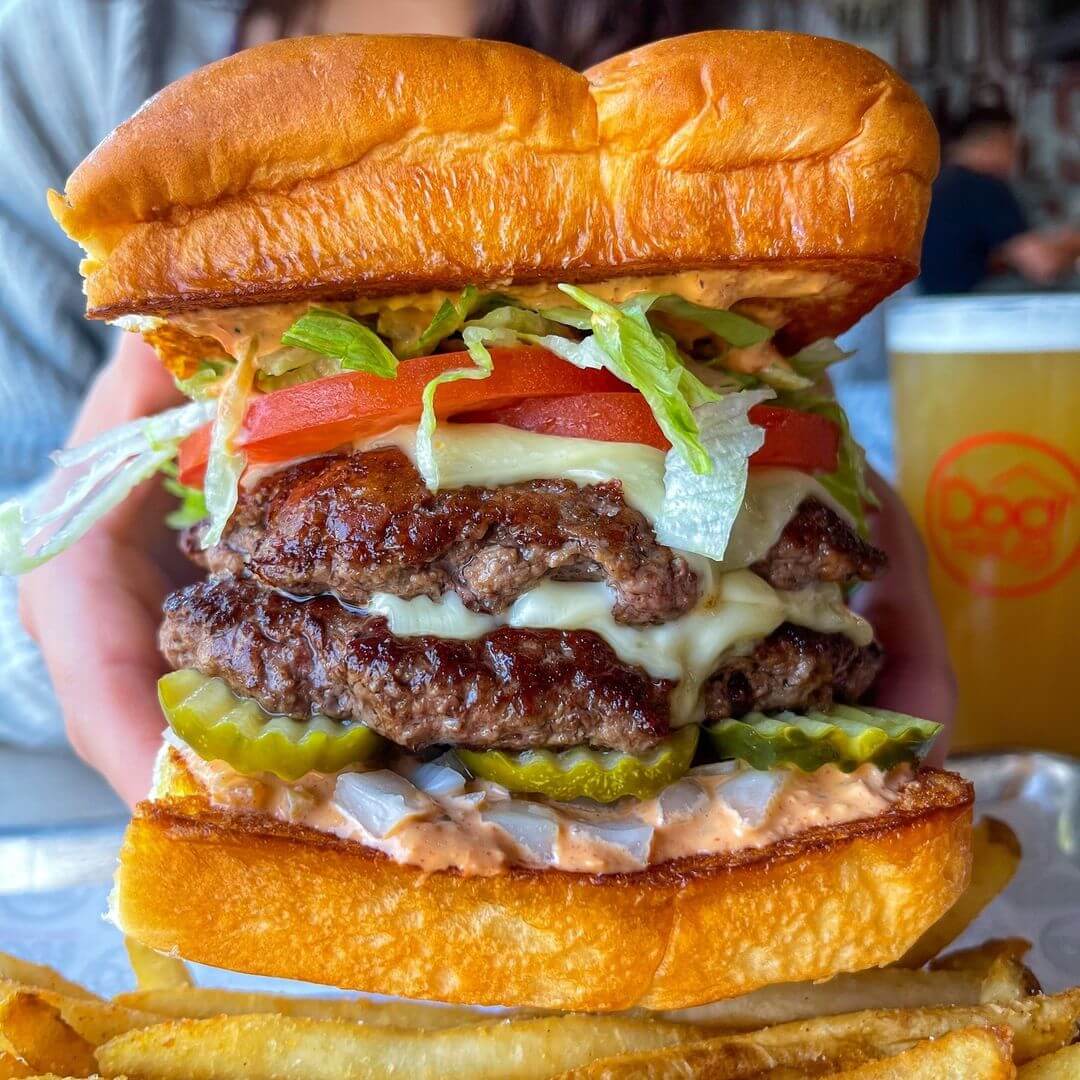 Close-up of a double cheeseburger with lettuce, tomato, pickles, onions, and sauce, served with a side of fries, held by a person. A drink is in the background.