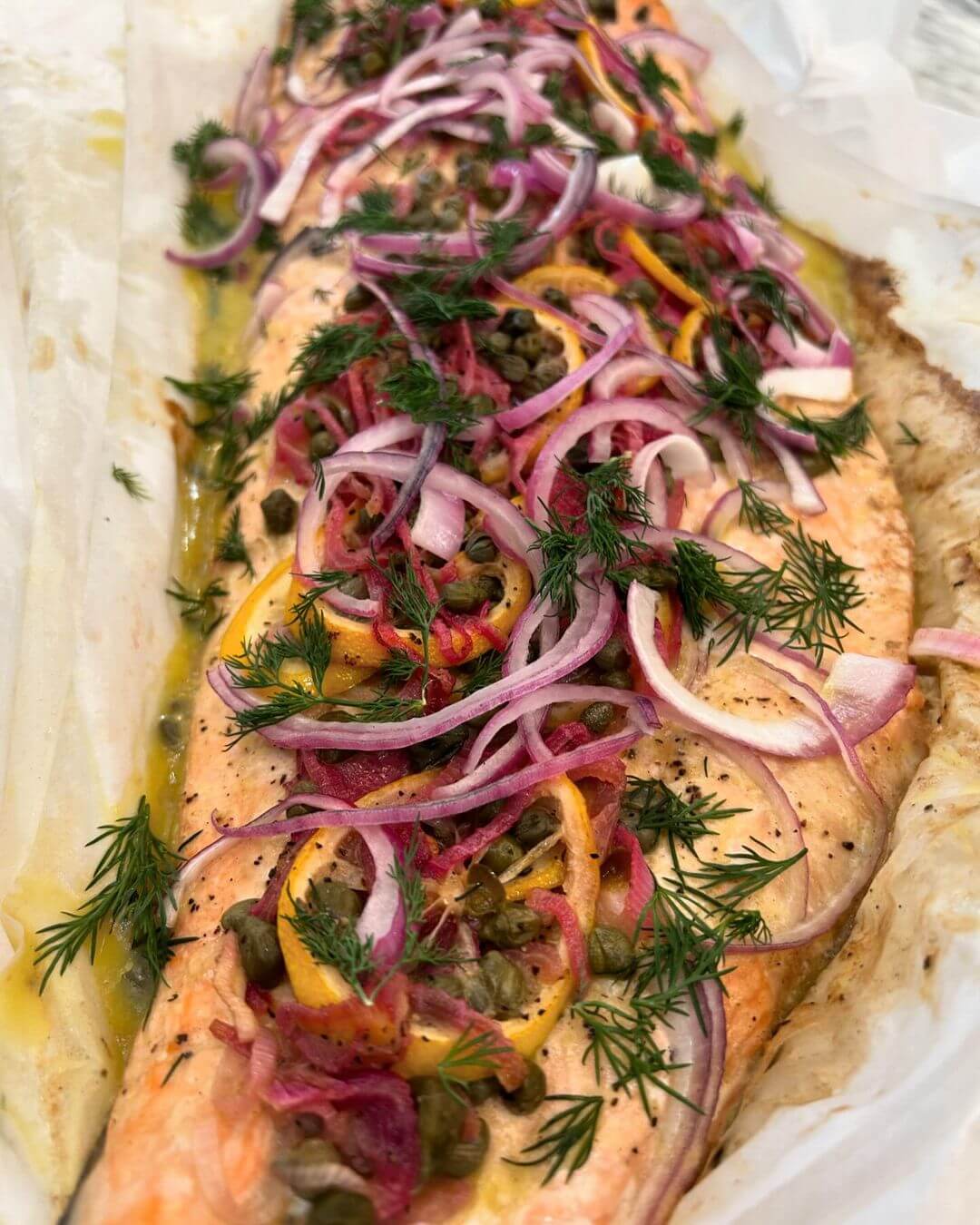 A roasted salmon fillet is garnished with sliced red onions, capers, lemon slices, and fresh dill. The dish is presented on parchment paper.