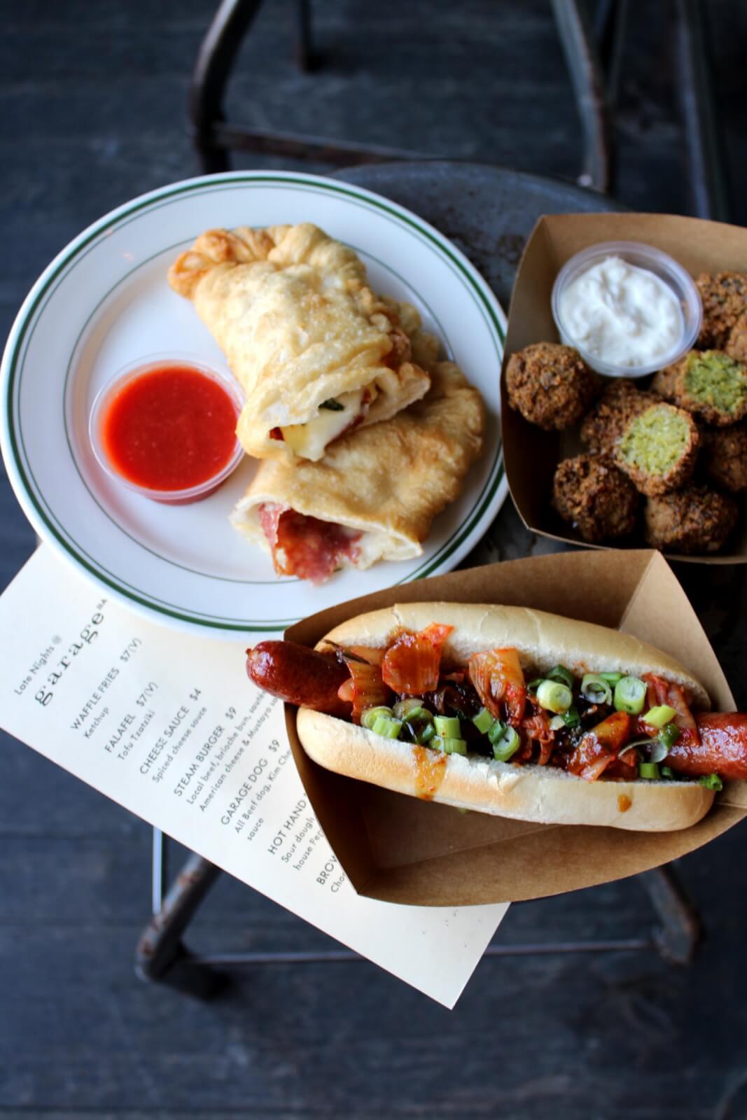 A plate with empanadas and red dipping sauce, a container of falafel balls with white sauce, and a hot dog topped with vegetables, placed on a table with a menu underneath.
