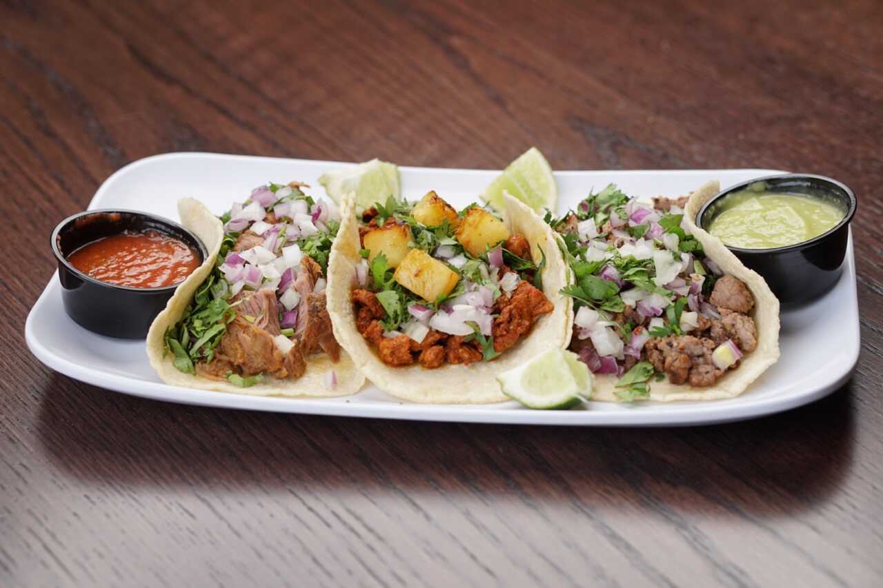 A plate with three tacos, each garnished with chopped onions, cilantro, and lime wedges, served with small bowls of red and green sauce on the side.