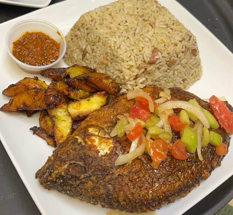 A plate with fried fish topped with vegetables, fried plantains, rice with bits of meat, and a small cup of sauce.