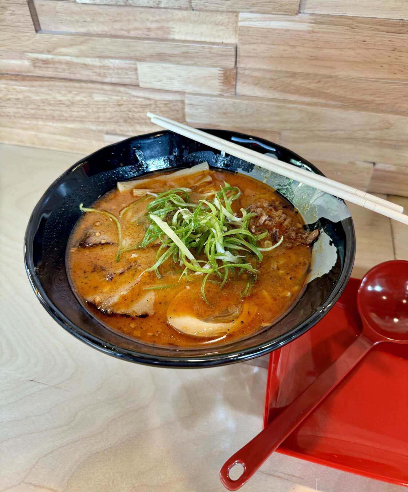 A bowl of ramen with sliced pork, green onions, and chopped vegetables in a rich, orange broth. There are chopsticks resting on the bowl and a red spoon on a red tray beside it.