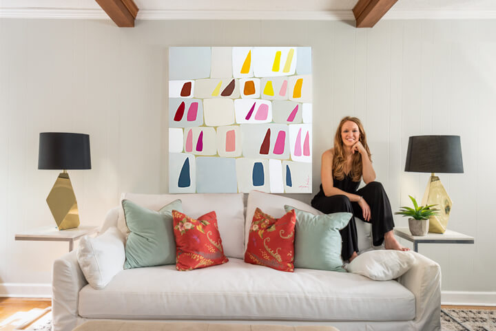 A woman dressed in black sits on the arm of a white sofa with colorful pillows. Above her hangs a large abstract painting. Two black lamps sit on tables on either side of the sofa, creating a chic FACE of Memphis ambiance.