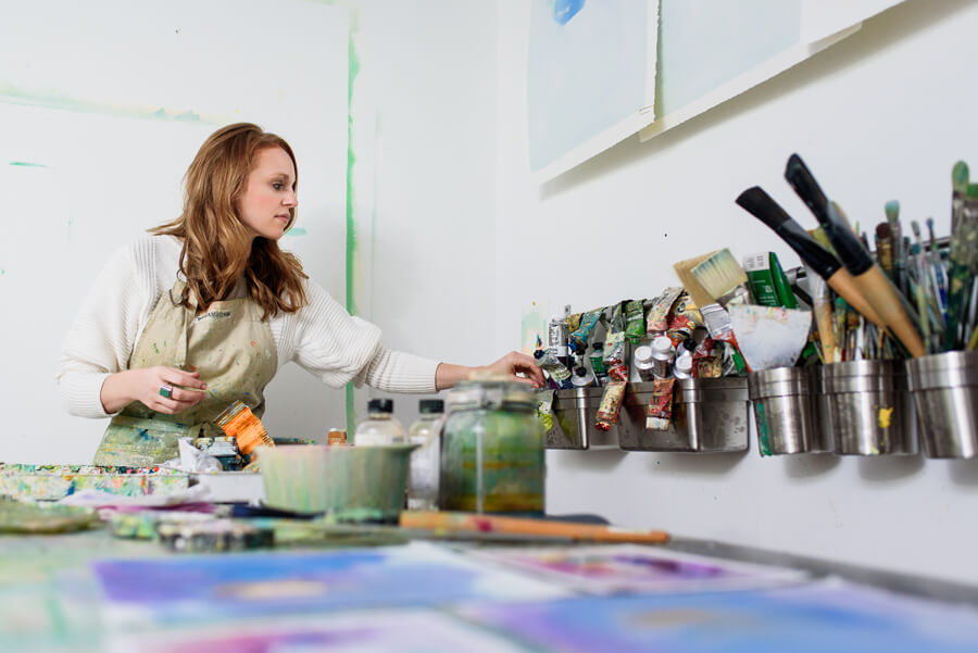 Hillary Butler is wearing an apron and selects a tube of paint from a wall-mounted rack in an art studio. Paint tubes, brushes, and art supplies are visible on the table and in containers.