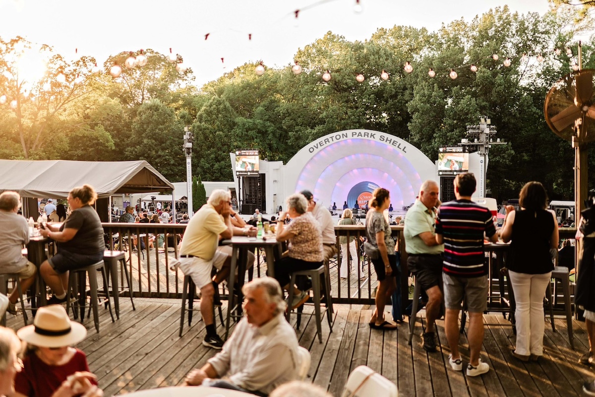 People sit and stand at outdoor tables on a wooden deck overlooking the Overton Park Shell stage, enjoying live performances amid trees in the background during events in June.