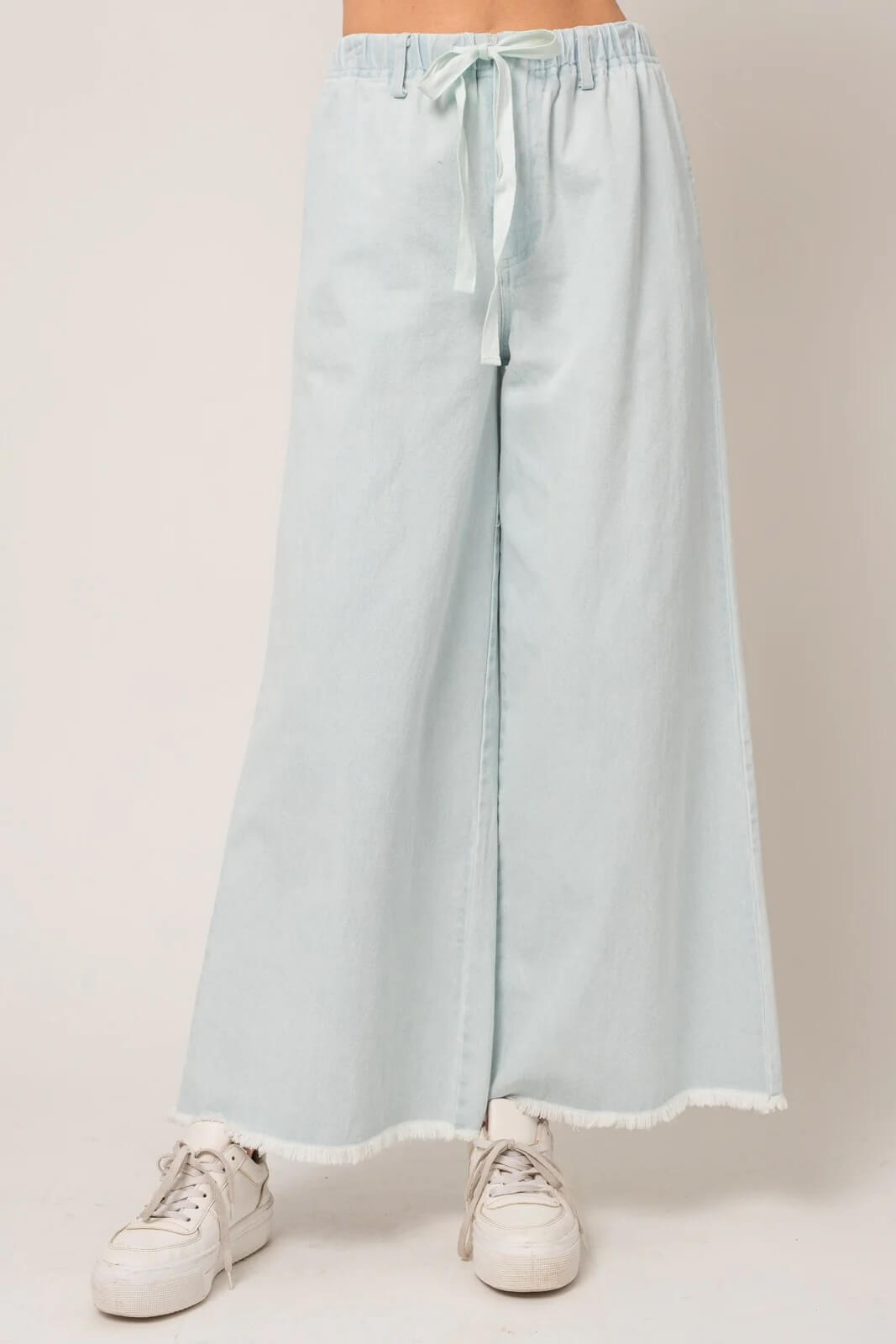 Light blue wide-leg trousers with a frayed hem and white drawstring, paired with white sneakers, displayed against a neutral Memphis background.