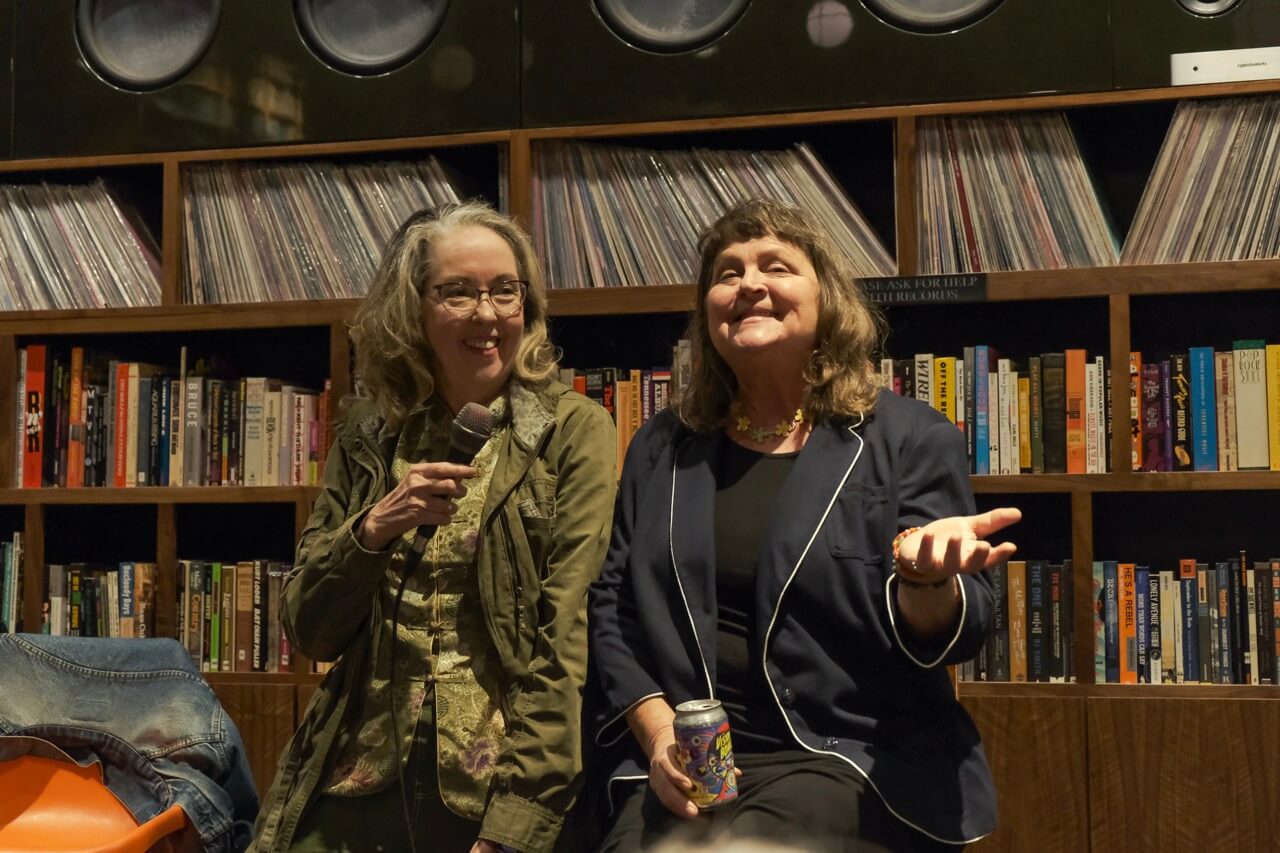 5 Local Experiences Every Memphian Should Try - 7 Two women are seated in front of a bookshelf filled with records and books. One holds a microphone, while the other holds a can. They appear to be speaking or engaging with an audience.