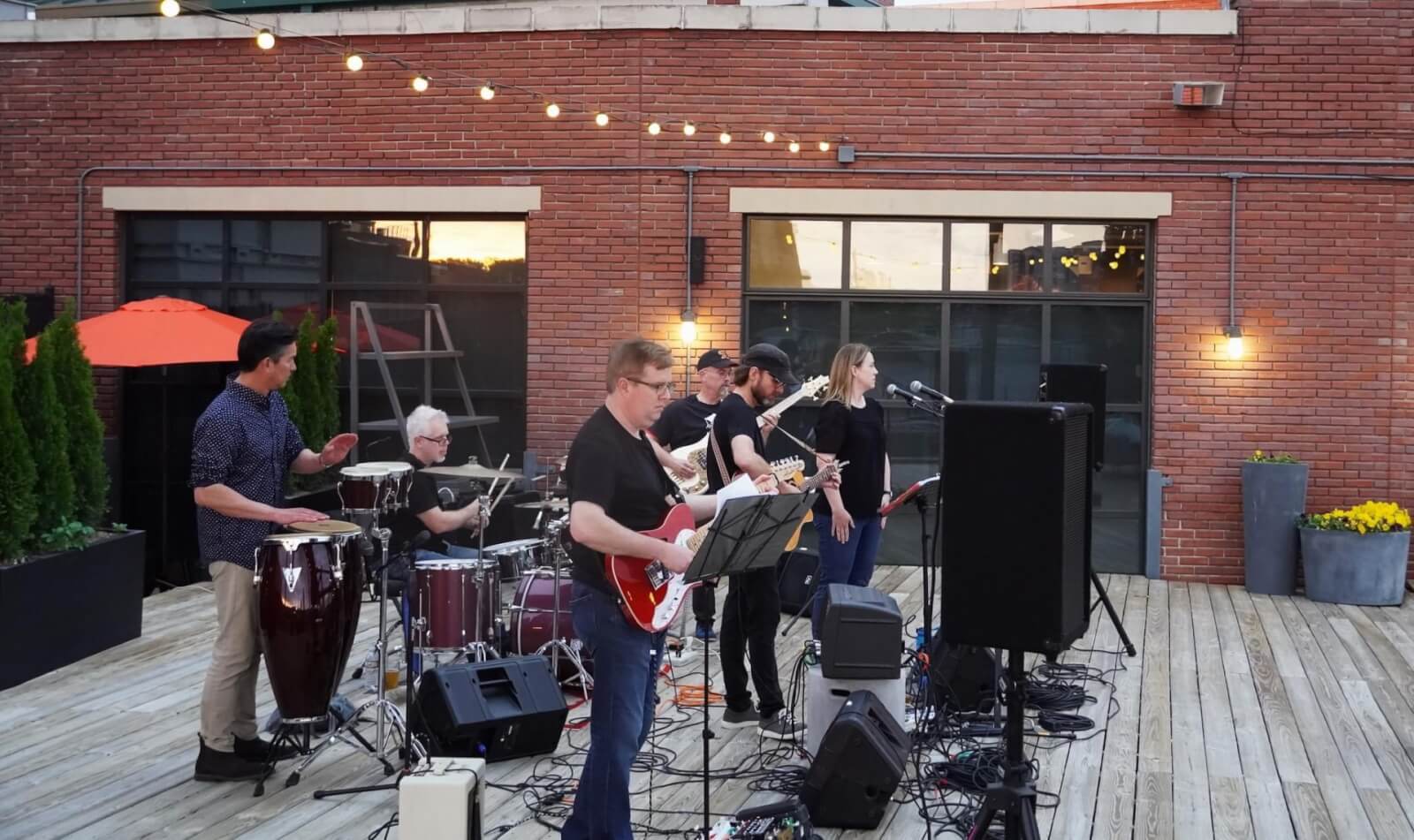 5 Local Experiences Every Memphian Should Try - 10 A band performs on a wooden deck in front of a brick building, with string lights overhead. Musicians play guitar, drums, and percussion, while one performer sings into a microphone.