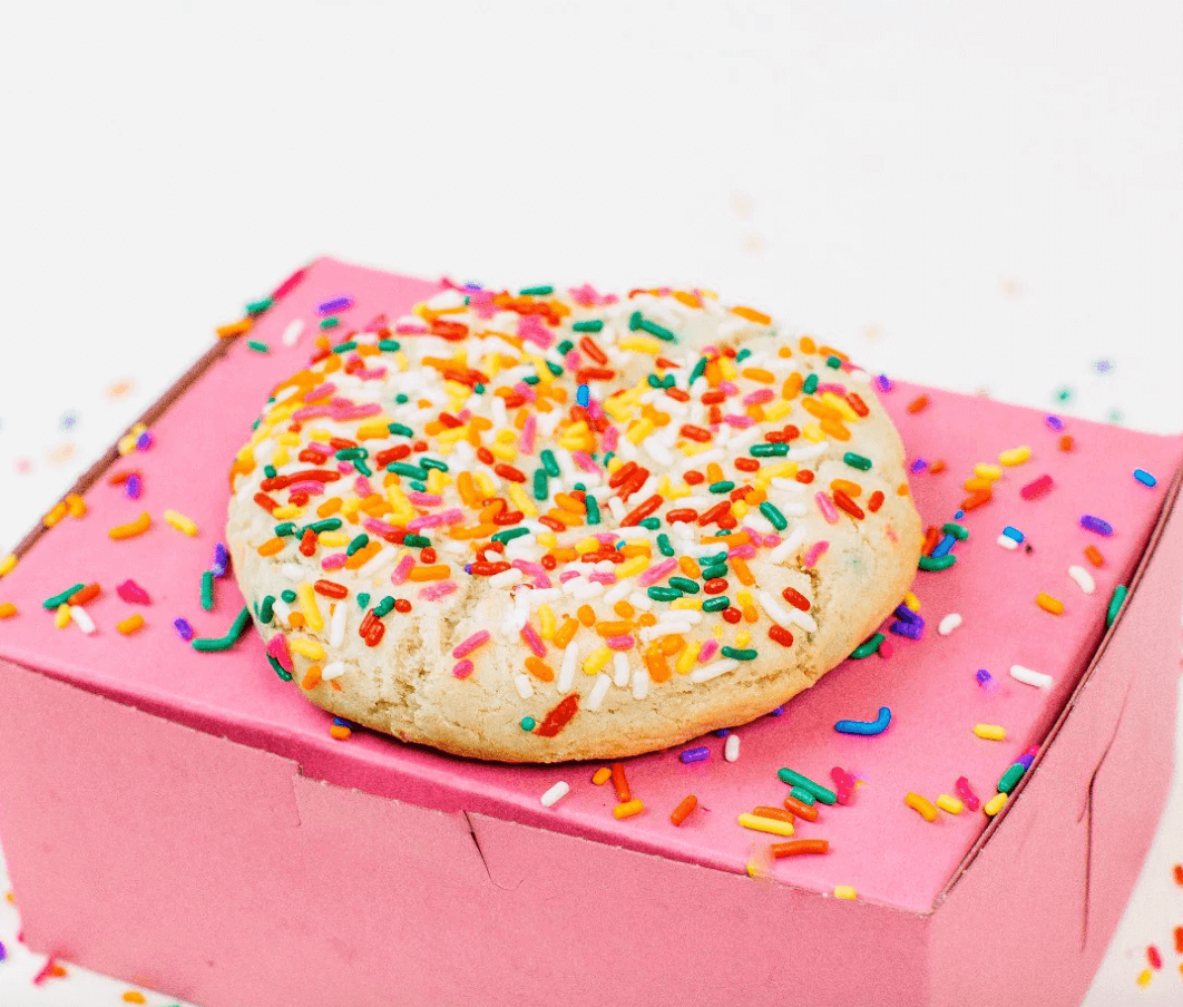 A colorful sprinkled cookie resting on a pink gift box against a white background.