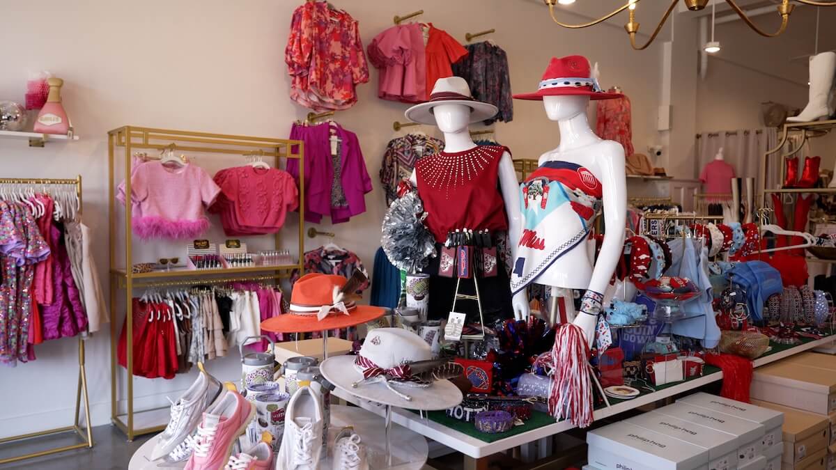 A boutique display featuring mannequins dressed in colorful, festive outfits with hats, surrounded by various apparel and accessories, including shoes, belts, and bags, against a neatly arranged background.