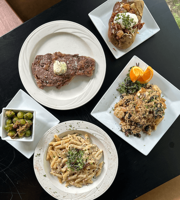 A table is set with a meal that includes a plate of penne pasta, a grilled steak with butter, a loaded baked potato, a serving of Brussels sprouts, and a dish of seasoned rice topped with orange slices.