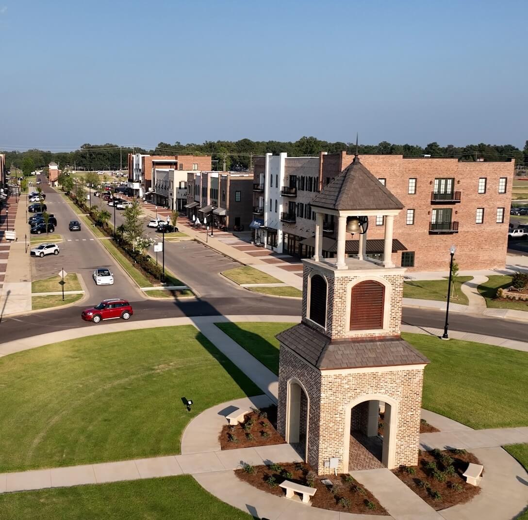 Aerial view of a clock tower on a green lawn with pathways, surrounded by a road, parked cars, and buildings in the background.