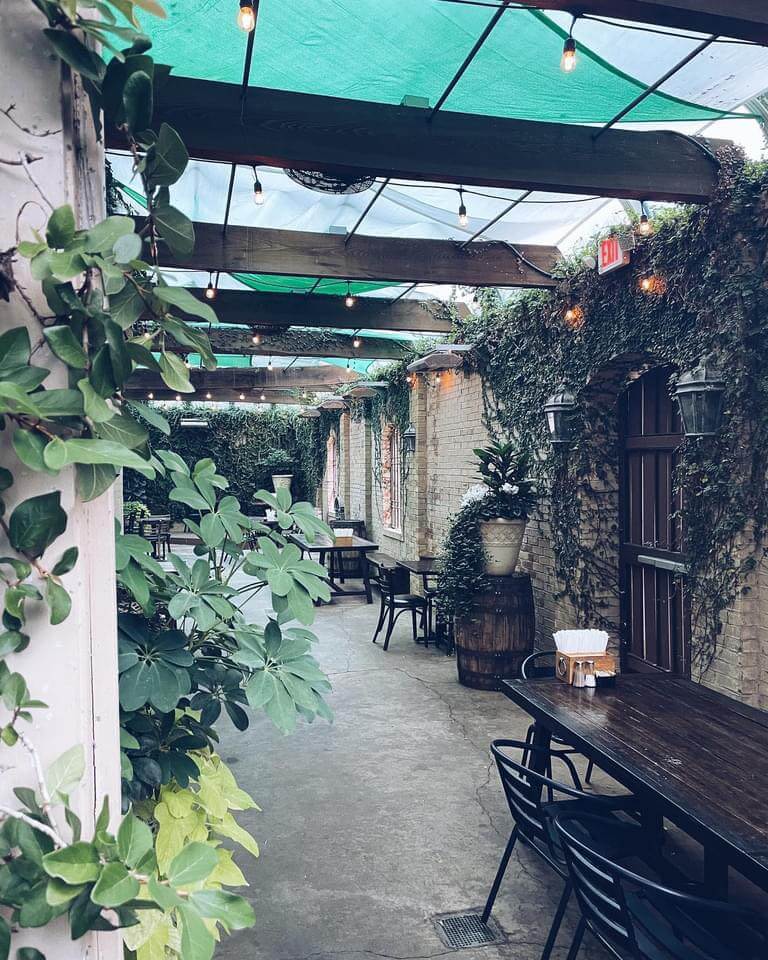 Outdoor patio with wooden tables and chairs, surrounded by green plants climbing the walls. Overhead, string lights hang beneath a green canopy. This Mississippi summer destination has a rustic, cozy ambiance perfect for warm evenings.