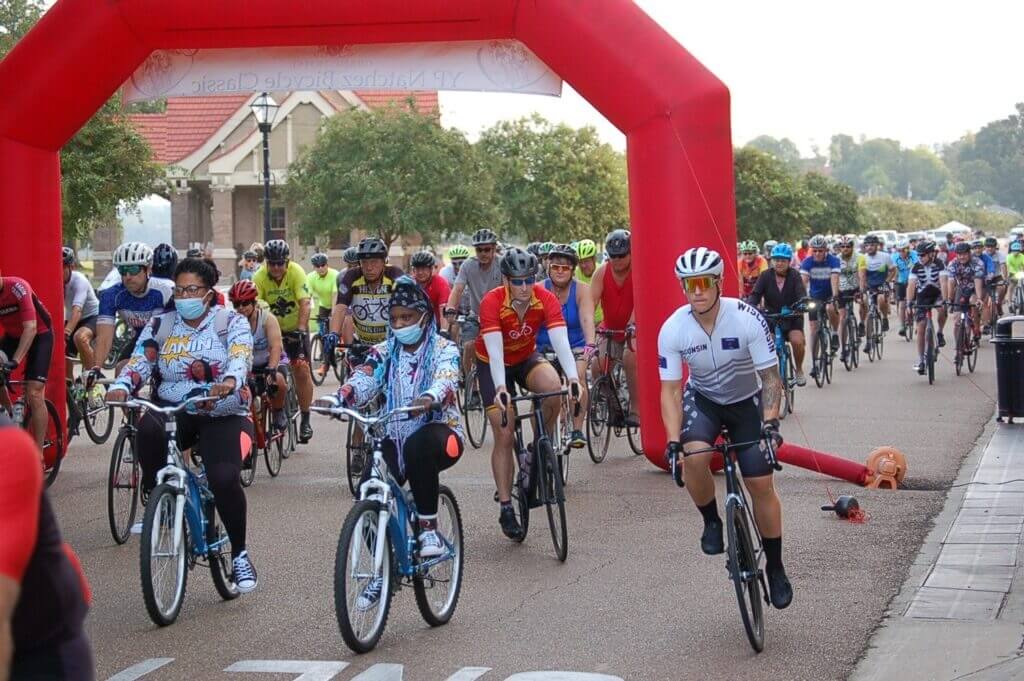 A group of cyclists in colorful gear participate in a cycling event, riding under a large red inflatable arch on a cloudy day.