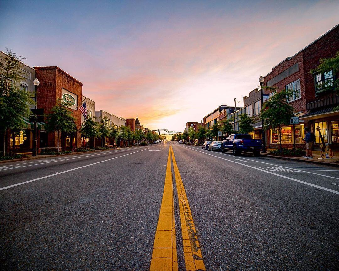 A main street in a town at sunset, featuring a clear road with a double yellow line, bordered by buildings, trees, and parked cars. The sky shows hues of orange, pink, and blue.