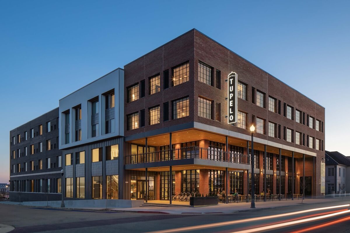 Four-story modern brick building with "Hotel Tupelo" sign illuminated on the corner, featuring large windows and an exterior patio, seen at dusk with light trails from passing cars.