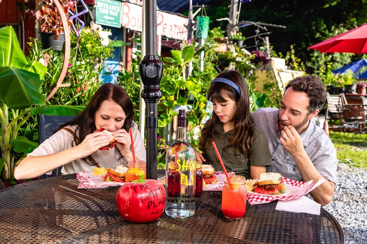 A woman, a child, and a man sit outdoors at a table, eating burgers. The table has a large tomato decoration.