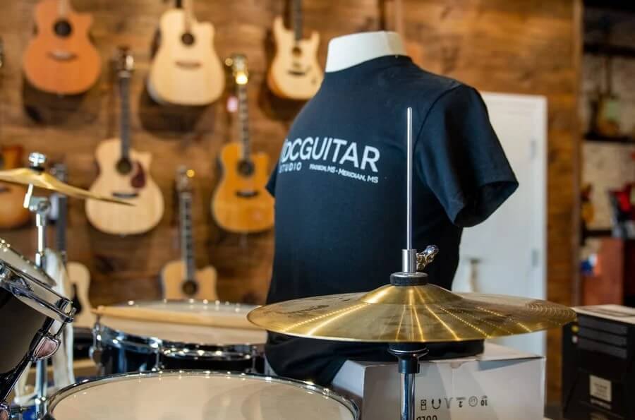 A drum set and cymbals in the foreground, with several acoustic guitars and a mannequin wearing a black T-shirt in the background inside a music store.