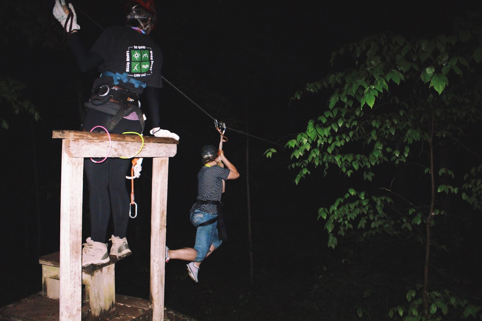 A person ziplining at night guided by an instructor on a wooden platform, surrounded by dark foliage.